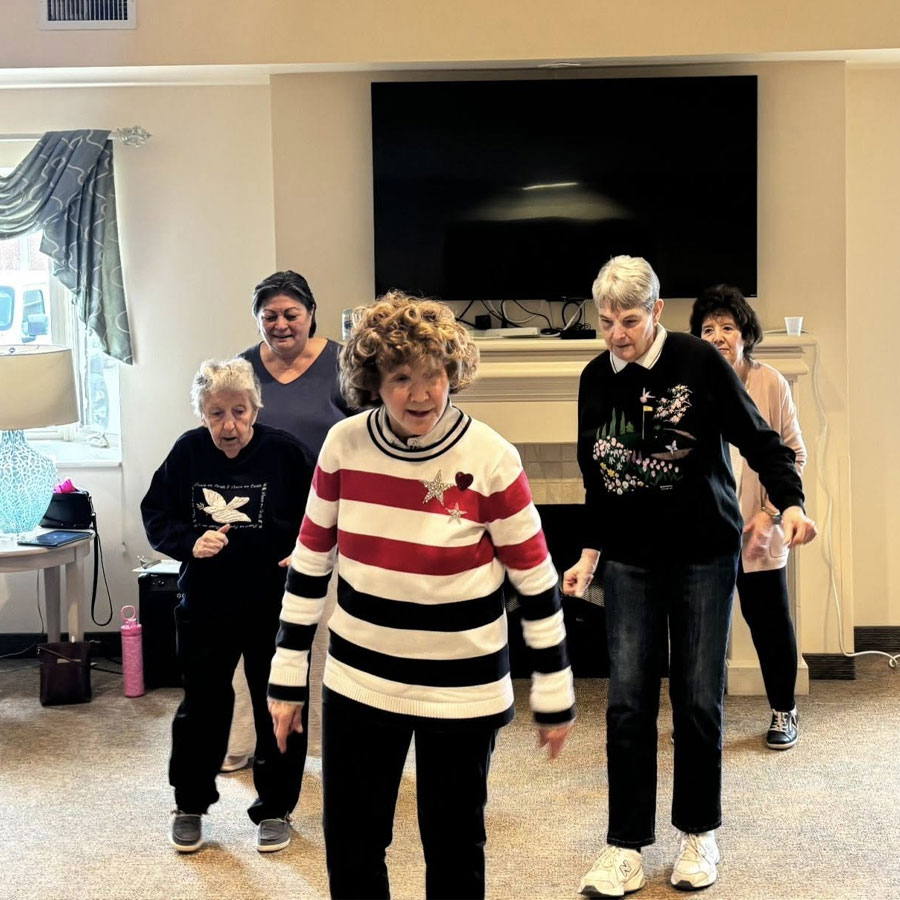 Five senior women doing a group dance in a lounge by a TV and fireplace; one wears a red-and-white striped sweater